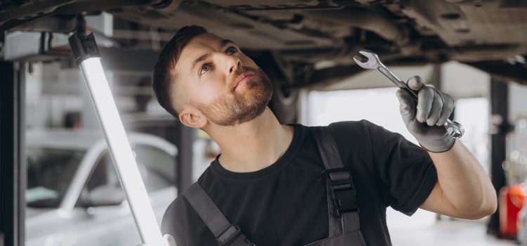 View from side of concentrated worker of car service checking running gear of automobile. Male mechanic in uniform and gloves standing under car, keeping lamp and looking for breakage